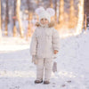 Enfant debout dans une forêt enneigée portant l'ensemble de ski écru avec moufles - Bébé Lova