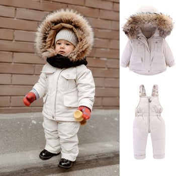 Bébé debout en extérieur portant l'ensemble de ski beige, avec aperçu de la doudoune et de la salopette - Bébé Lova