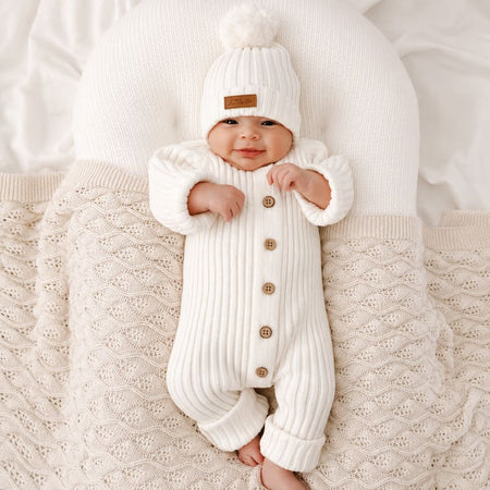 Combinaison tricot bébé blanche portée par un bébé souriant avec bonnet à pompon - Bébé Lova