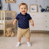 Bébé garçon souriant debout dans une chambre portant l'ensemble polo marine à ancres et short beige - Bébé Lova