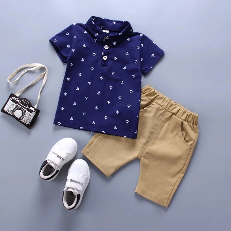 Ensemble 2 pièces garçon polo marine à ancres et short beige présenté à plat avec sac appareil photo et baskets - Bébé Lova