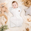 Bébé allongé dans une gigoteuse swaddle grise rayée avec décor cocooning et peluche ours - Bébé Lova