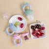 Grignoteurs de fruits en silicone vert, bleu et rose présentés avec des raisins et des cerises frais - Bébé Lova