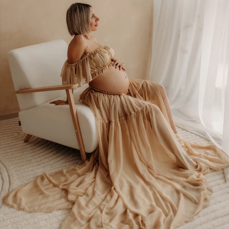 Femme enceinte portant une robe longue beige fluide assise dans un fauteuil élégant pour séance photo grossesse - Bébé Lova