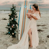 Jeune maman portant son bébé sur la plage avec robe de maternité fluide et planche de surf - Bébé Lova