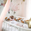 Bébé allongé dans un lit à barreaux avec un tour de lit tressé blanc, caramel et gris dans une chambre aux tons roses - Bébé Lova