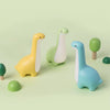 Les trois coloris de la veilleuse dinosaure (jaune, vert et bleu) dans un décor végétal - Bébé Lova